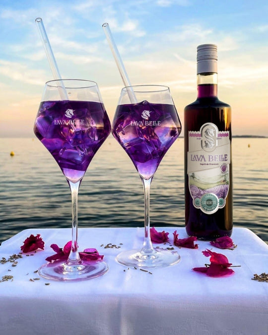 Two Lav'a Belle glasses of purple spritz with glass straws and petals next to a bottle by the sea at dusk
