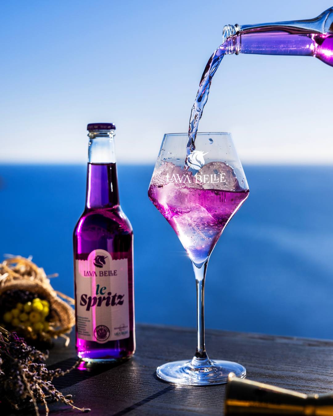 Bottle of Lav'a Belle le Spritz against a blue sea, purple spritz being poured into a glass with ice beside it