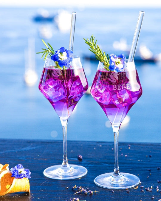 Two Lav'a Belle Signature apéritif glasses with engraved logo, filled with purple spritz against a sea backdrop