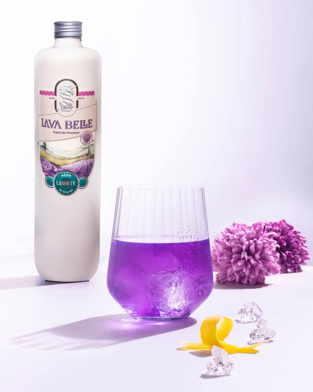 Lav'a Belle Liberté: white bottle behind a glass of purple drink on ice, with lemon zest and a purple blossom