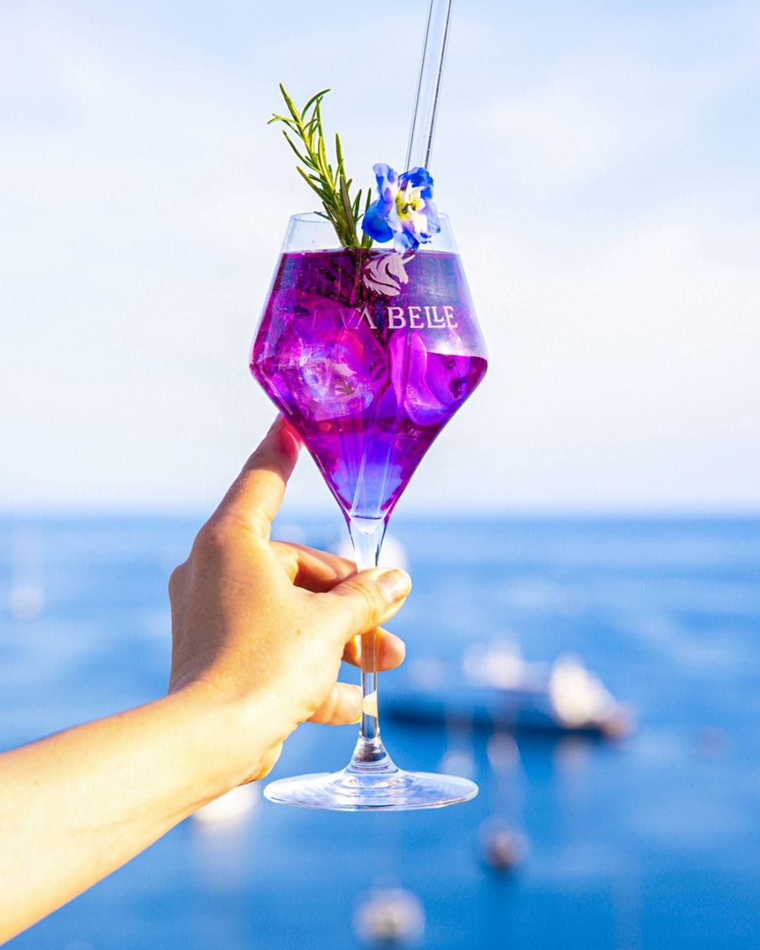Hand holding a Lav'a Belle glass of purple spritz with rosemary and a blue blossom against sea and sky