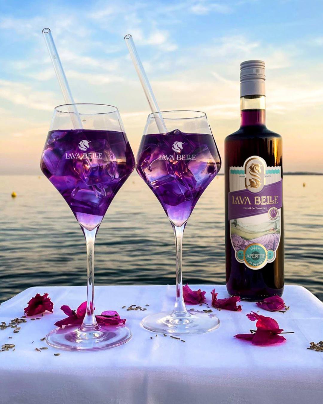 Two Lav'a Belle glasses of purple spritz with glass straws next to a bottle of Apéritif by the sea in evening light