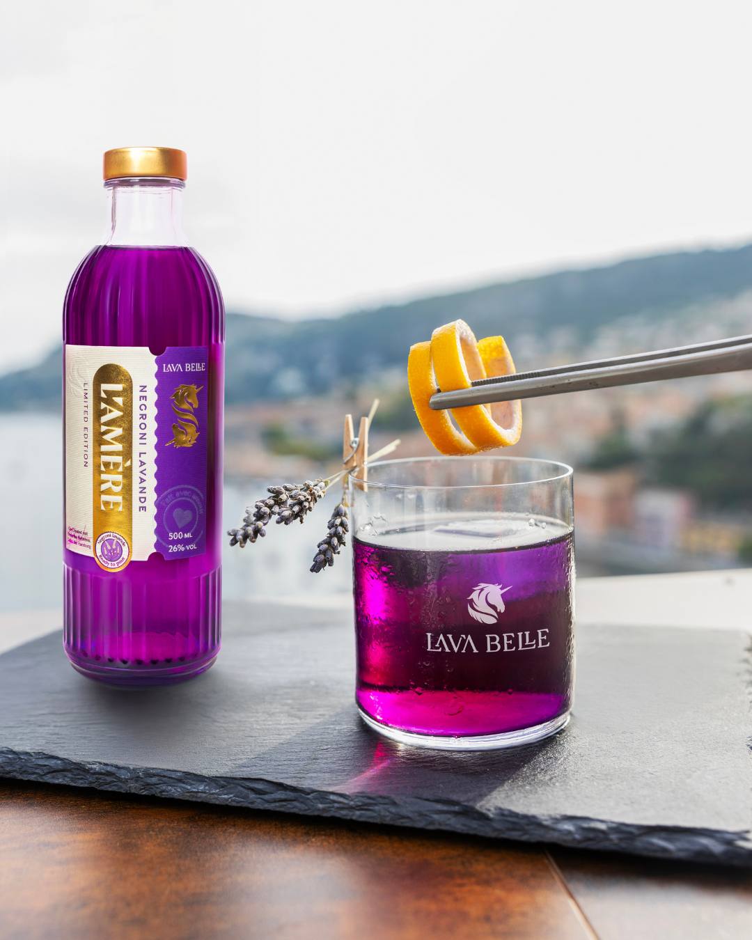 Bottle of L'Amère Negroni Lavande next to a Lav'a Belle glass of purple drink, orange zest and lavender against a coastal view