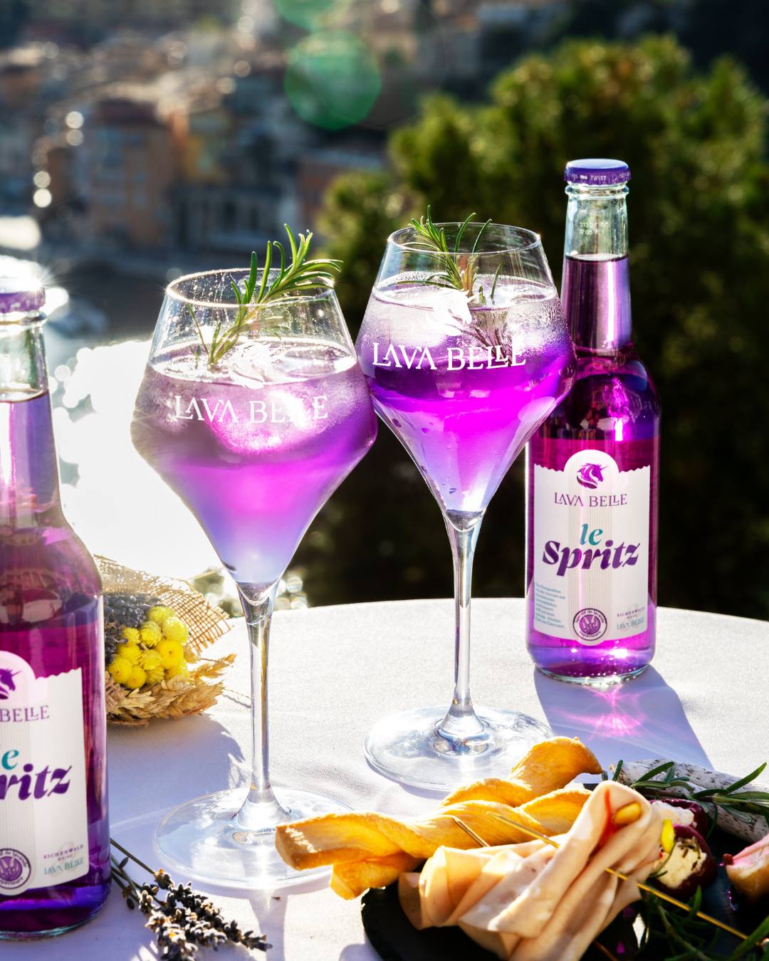 Two le Spritz bottles and Lav'a Belle glasses with rosemary on a laid table with snacks above a coastal town
