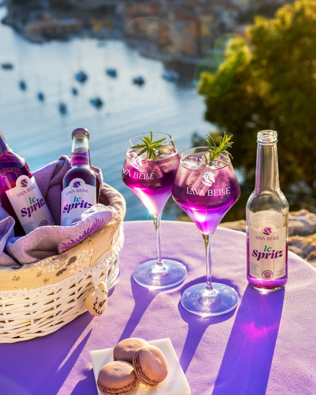 Picnic with Lav'a Belle le Spritz: bottles in a basket, two glasses with rosemary and macarons above a bay