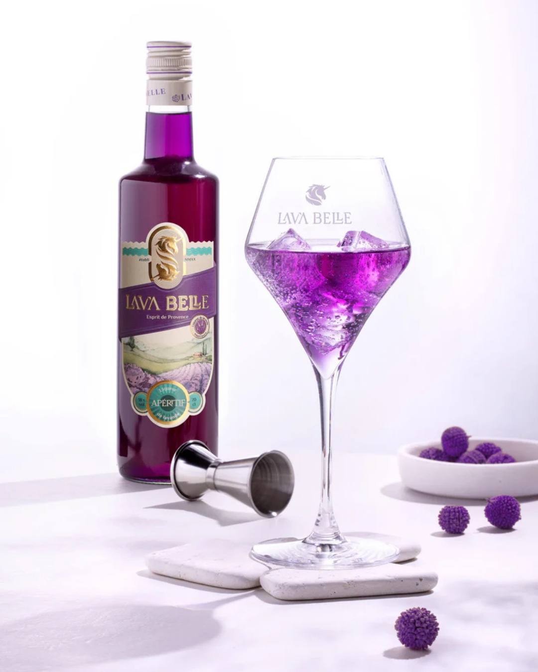 Bottle of Lav'a Belle Apéritif next to a glass of purple drink on ice, with a jigger and purple decorative baubles