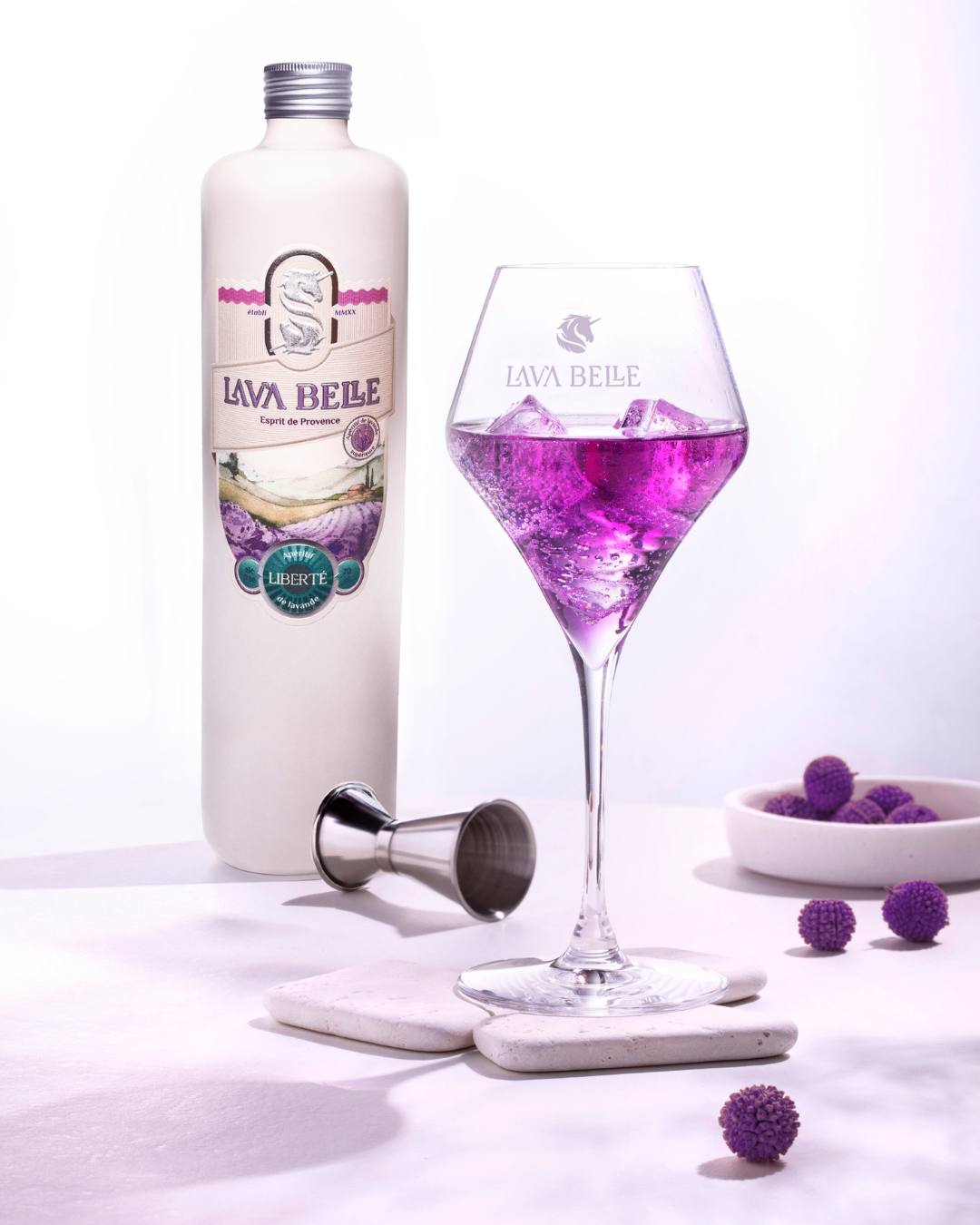 White bottle of Lav'a Belle Liberté next to a glass of purple drink on ice, with a jigger and purple decorative baubles